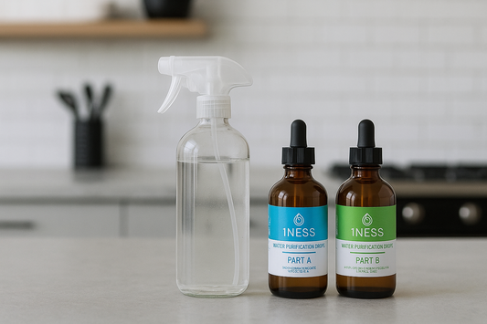 Glass spray bottle with water and two brown dropper bottles labeled "1NESS Water Purification Drops Part A and Part B" on a kitchen counter for making chlorine dioxide disinfectant solution.